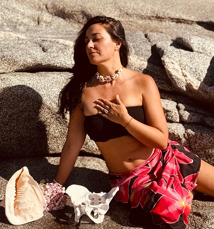 Bernardita Bordagorry está sentada sobre unas rocas con los ojos cerrados, vestida con un top negro de tirantes, un pareo de flores rojas y un collar de conchas. A su lado hay una caracola, un modelo de hueso pélvico y flores.