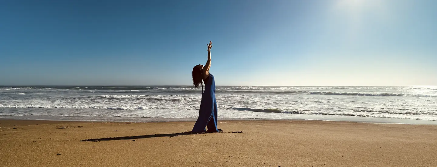Bernardita Bordagorry está de pie en una playa de arena con un vestido largo, estirando los brazos hacia el cielo azul claro con el océano a su lado.