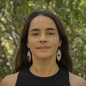 Josefina López de pelo largo y oscuro, con un top negro sin mangas y pendientes ovalados, está de pie al aire libre delante de un follaje verde.
