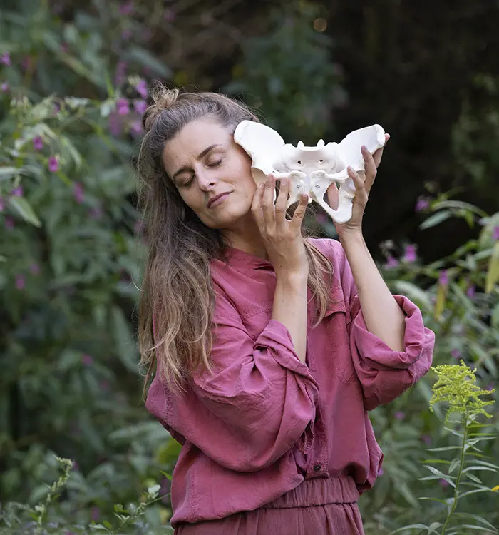 Magdalena Haftner de pelo largo, se sienta descalza en un taburete al aire libre, rodeada de denso follaje verde y plantas.