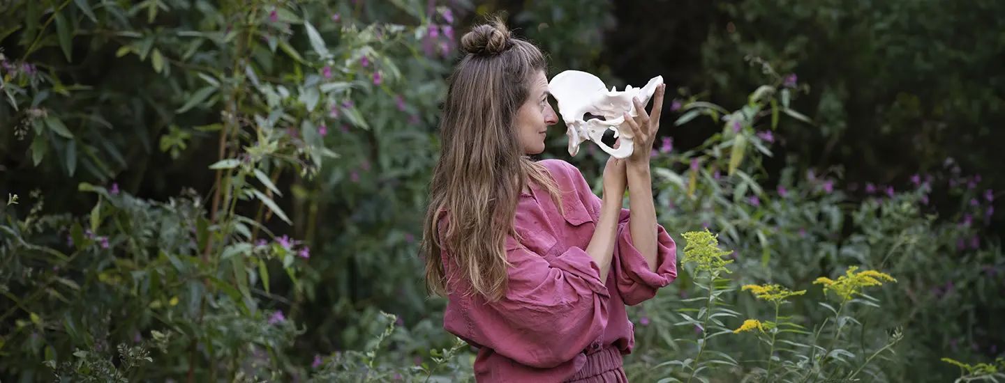 Magdalena Haftner está agazapada sobre un tronco caído en un bosque iluminado por el sol, rodeada de frondosa vegetación y altos árboles.