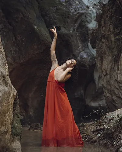 Una mujer con un vestido rojo vaporoso, posa elegantemente con un brazo levantado y la cabeza inclinada hacia un lado en aguas poco profundas, entre acantilados rocosos.