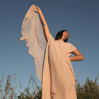 Una mujer está de pie, con un vestido ligero y vaporoso, al aire libre, con un brazo levantado, sosteniendo una tela contra un cielo azul claro. En primer plano, hierbas altas y plantas se mecen suavemente.