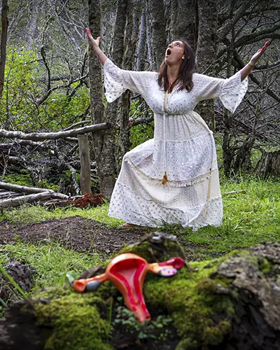 Mujer está de pie en un claro del bosque, con los brazos levantados y la boca abierta en señal de expresión; un modelo de útero descansa sobre musgo en primer plano, evocando temas de naturaleza y feminidad.