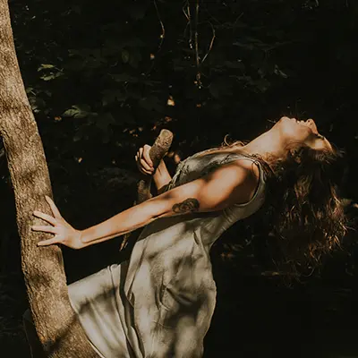 Mujer con un vestido claro, se apoya en un árbol de un bosque, sujeta una rama y mira hacia arriba, con el rostro parcialmente iluminado por la luz del sol.