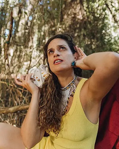 Clara Davaar, con un vestido amarillo sin mangas, está sentada al aire libre sosteniendo una concha marina junto a la oreja y mirando hacia arriba, rodeada de árboles y de la luz del sol.