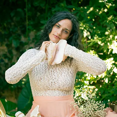 Clara Davaar, con un top de encaje y falda melocotón, está de pie al aire libre con vegetación de fondo, sujetando una gran caracola al cuello.