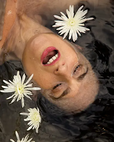 Clara Davaar, con un pintalabios rosa brillante, está parcialmente sumergida en agua oscura, rodeada de cuatro flores blancas que flotan cerca.